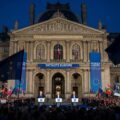 A photo-realistic image captures a 'Patriots Europe' rally in France, with a grand, classical French building as the backdrop. Marine Le Pen and Viktor Orban are prominently positioned, addressing a large crowd. The dramatic lighting emphasizes both the architecture and the speakers, reflecting a national conservative vision that stands in opposition to the EU."