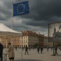 Photo-realistic image of a peaceful election scene in a city square in Poland, with citizens voting under a white canopy near Polish and EU flags. Dark storm clouds gather overhead, symbolizing anticipated unrest. In the background, a billboard displays ominous Russian interference imagery, suggesting a threat that never materialized. A police officer stands watch nearby.