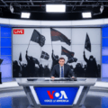 A 16:10 photo-realistic image of a Voice of America broadcast studio with an anchor at a desk, facing the camera. Behind the anchor, a large screen displays a live feed of a stylized, somewhat blurred rally featuring aggressive figures holding dark flags, visually representing the kind of far-right content often associated with figures like Kari Lake or networks such as OAN.