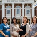 Four pregnant women in front of the heritage foundation showing the impact of global national conservative alliance, pronatalist policies.