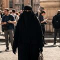 A photorealistic image depicts a woman wearing a black niqab and abaya, with only her eyes visible, walking through a bustling public square in Italy.